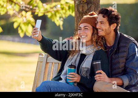 Smiling Couple Taking Selfie With Mobile Phone Sitting On Bench In Autumn Park Stock Photo