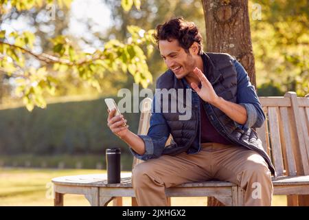 Smiling Man Sitting On Bench Making Video Call In Autumn Park Using Mobile Phone Stock Photo