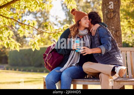 Hugging Couple Sitting On Bench In Autumn Park With Reusable Drinks Cups Stock Photo