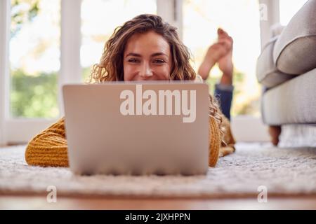 Woman In Warm Jumper Lying On Floor At Home Using Laptop Stock Photo