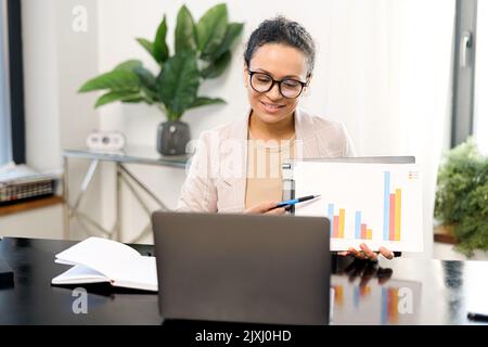 Businesswoman involved conference, tutor or business coach leading webinar. Middle aged female employee during virtual meeting, showing graphs into webcam of the laptop Stock Photo