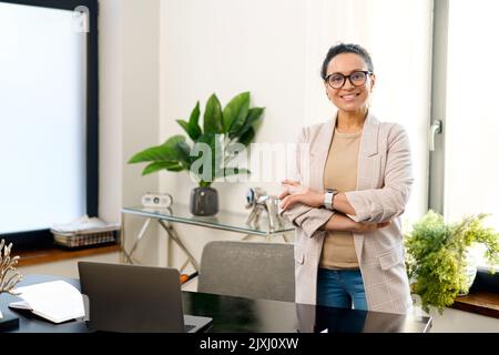 Cheerful woman entrepreneur stands with arms crossed in the contemporary office, freelancer woman in smart casual wear looks at the camera with arms crossed, small business owner Stock Photo