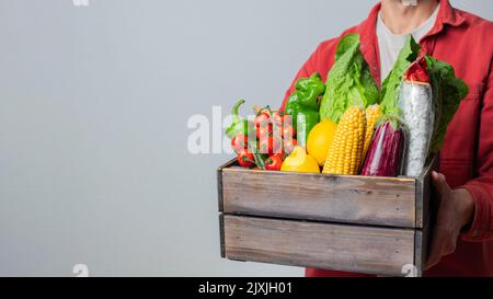 Delivery Concept -Young Caucasian delivery man carrying wooden box of grocery vegetables and fruits from store. Isolated on Grey studio Background Stock Photo
