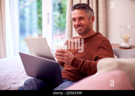 Mature Man At Home Sitting On Sofa With Laptop Looking Through Personal Finance Documents Stock Photo