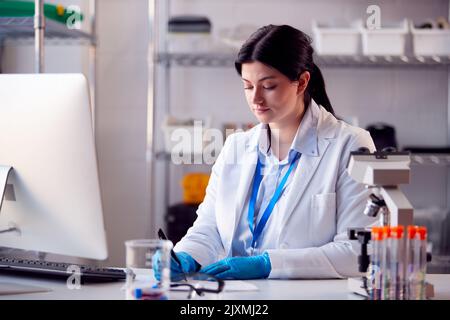 Female Lab Worker Wearing White Coat Recording Test Results On Computer Stock Photo