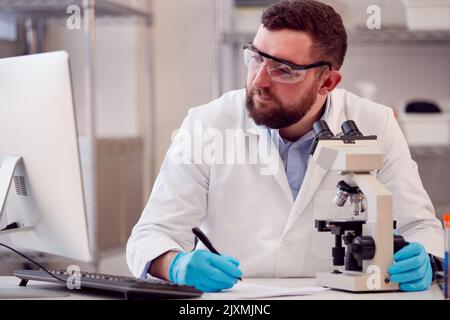 Male Lab Worker Wearing White Coat Recording Test Results On Computer Stock Photo