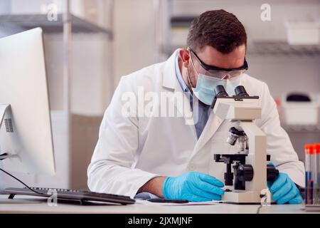 Male And Female Lab Workers Conducting Research Using Microscope And Recording Result On Computer Stock Photo