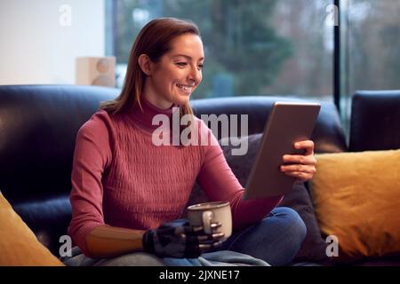 Woman With Prosthetic Arm And Hand At Home Using Digital Tablet Drinking Cup Of Coffee Stock Photo