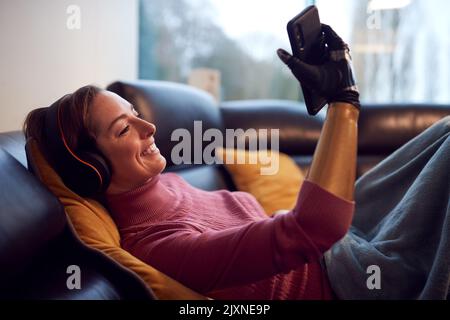 Woman With Prosthetic Arm Wearing Wireless Headphones Listening To Music On Mobile Phone On Sofa Stock Photo