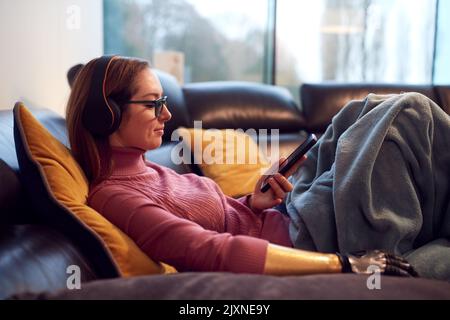 Woman With Prosthetic Arm Wearing Wireless Headphones Listening To Music On Mobile Phone On Sofa Stock Photo