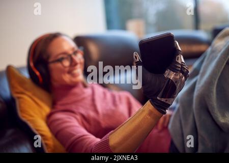 Woman With Prosthetic Arm Wearing Wireless Headphones Listening To Music On Mobile Phone On Sofa Stock Photo