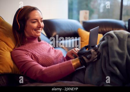 Woman With Prosthetic Arm Wearing Wireless Headphones And Watching Movie On Laptop On Sofa At Home Stock Photo