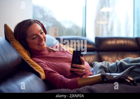 Woman With Prosthetic Arm Wearing Wireless Headphones Listening To Music On Mobile Phone On Sofa Stock Photo