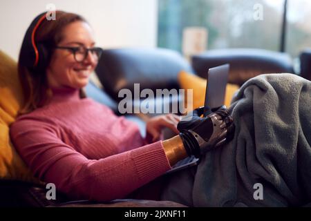 Woman With Prosthetic Arm Wearing Wireless Headphones And Working On Laptop On Sofa At Home Stock Photo