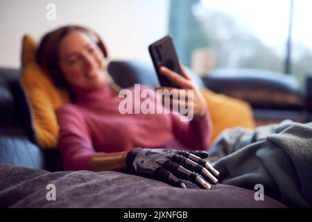 Woman With Prosthetic Arm Wearing Wireless Headphones Listening To Music On Mobile Phone On Sofa Stock Photo