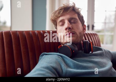 Man With Wireless Headphones Around Neck Relaxing On Armchair At Home With Closed Eyes Stock Photo