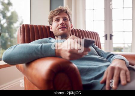 Man Sitting Relaxing On Armchair At Home Watching TV Using Remote Control Stock Photo