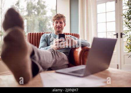 Man Relaxing In Armchair With Feet Up At Home Using Laptop And Mobile Phone Stock Photo