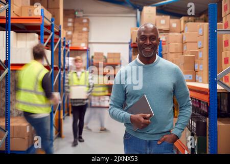Portrait Of Manager With Digital Tablet In Busy Modern Warehouse With Staff In Background Stock Photo