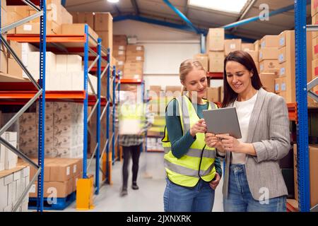 Manager Using Digital Tablet With Female Intern Inside Busy Warehouse Facility Stock Photo