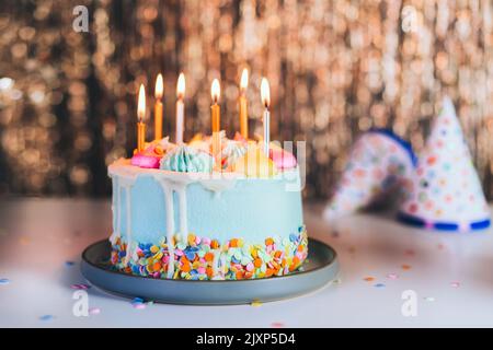 Colorful birthday cake with sprinkles and burning candles and festive caps on the sparkling gold tinsel background. Festive birthday celebration, part Stock Photo