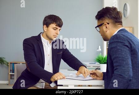 Two business people in office sign contract sitting at desk, manager gives document to his client. Stock Photo