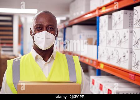 Portrait Of Male Worker Wearing PPE Face Mask Holding Box Inside Warehouse Stock Photo