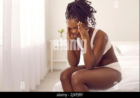 Tired stressed young black woman who is suffering from headache sitting on bed at home Stock Photo