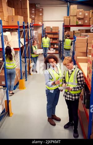 Male Intern With Team Leader Looking At Clipboard Inside Busy Warehouse Facility Stock Photo