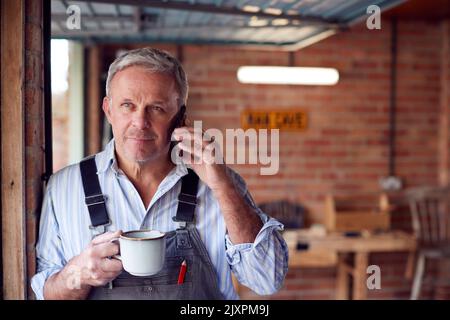 Mature Male Wearing Overalls Using Mobile Phone In Garage Workshop With Hot Drink Stock Photo