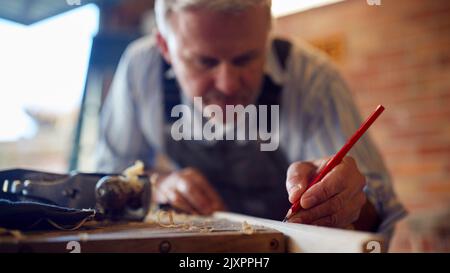 Mature Male Carpenter In Garage Workshop Marking Wood With Pencil Stock Photo