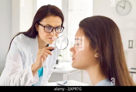 Caucasian woman doctor examines skin of female patient through magnifying glass in cosmetic clinic Stock Photo