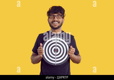 Portrait of Indian guy holding darts board with happy and confident face on orange background. Stock Photo