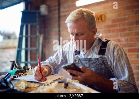 Mature Male Carpenter In Garage Workshop Working On Design Holding Mobile Phone Stock Photo