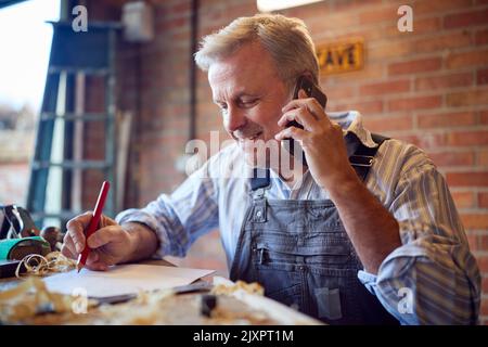 Mature Male Carpenter In Garage Workshop Working On Design And Talking On Mobile Phone Stock Photo