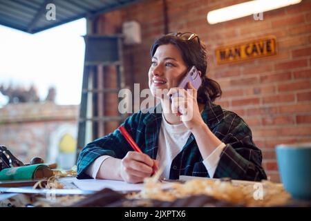 Young Female Carpenter In Garage Workshop Working On Design And Talking On Mobile Phone Stock Photo