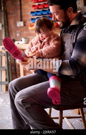 Father Helping Down Syndrome Daughter To Put On Wellington Boots In Garage Workshop At Home Together Stock Photo