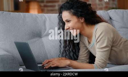 Side view happy user woman lying on couch sofa at home living room typing laptop keyboard working online ordering delivery from online shop computer Stock Photo
