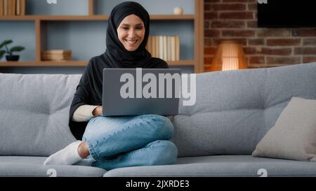 Muslim Islamic woman in hijab sitting at home living room relaxing on sofa couch using laptop online website ordering booking in internet shop working Stock Photo