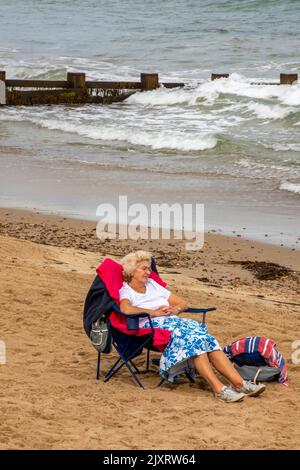 older lady falling asleep in a deckchair on a sandy beach at swanage, old lady sitting in a folding chair on a sandy beach on a summers day in dorset. Stock Photo