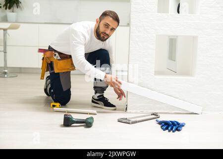 A man installs a floor skirting board. Fixing the plastic skirting board with screws to the wall. Home renovation. Stock Photo