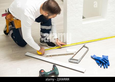 A man installs a floor skirting board. Fixing the plastic skirting board with screws to the wall. Home renovation. Stock Photo