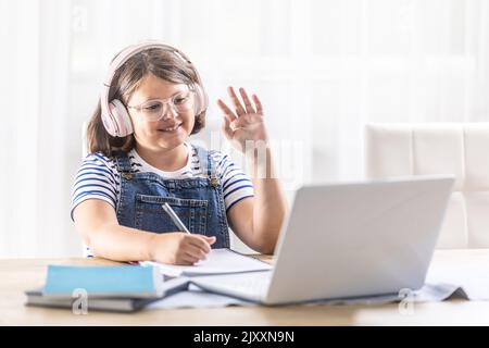 Chubby schoolgirl in glasses and headphones waves at her schoolmates on the computer during distant studies. Stock Photo