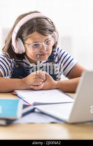 Hard-studying girl in glasses wears headphones and chews on pencil in her hand looking into the pc. Stock Photo
