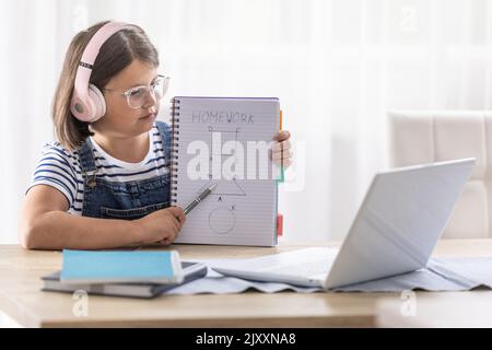 Schoolgirl in glasses and headphones shows her homework to the teacher during an online class via computer. Stock Photo
