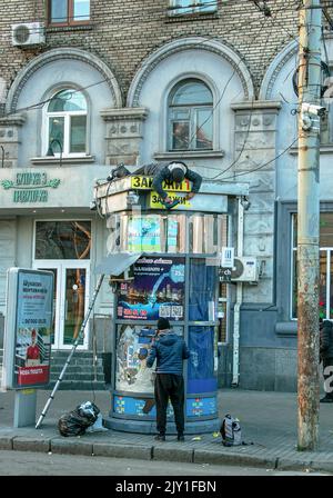 Dnipropetrovsk, Ukraine - 11.26.2021: A worker on the roof of a kiosk installs lighting devices with an electric drill. The worker lies on the roof of Stock Photo