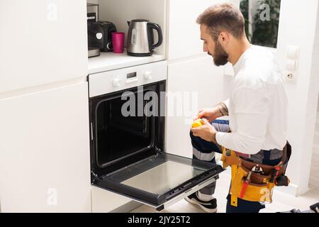 repairman examining oven with screwdriver in kitchen with tool case Stock Photo