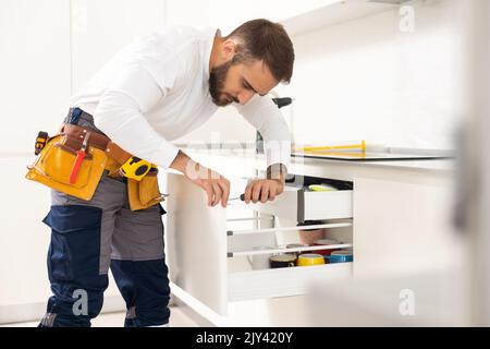Repairman In Overalls Repairing Cabinet Hinge In Kitchen Stock Photo