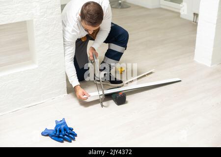 A man installs a floor skirting board. Fixing the plastic skirting board with screws to the wall. Home renovation. Stock Photo