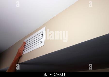 A left caucasian adult hand held up to a closed small air vent on a beige wall near a white ceiling. Hand on metal rectangle furnace closed vent grid Stock Photo